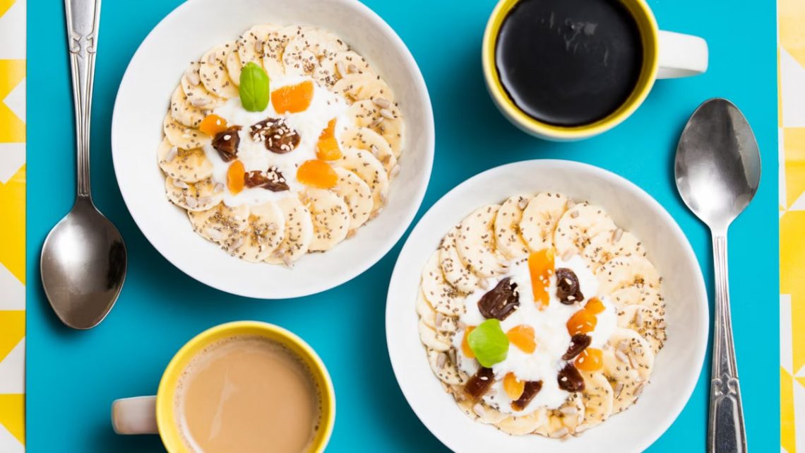 Top view of two breakfast bowls with bananas, yogurt, and coffee cups on a vibrant background.